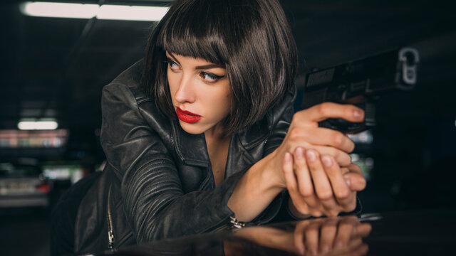 Beautiful brunette sexy spy agent (killer or police) woman in leather jacket and jeans with a gun in her hand running after someone, to catch him on parking