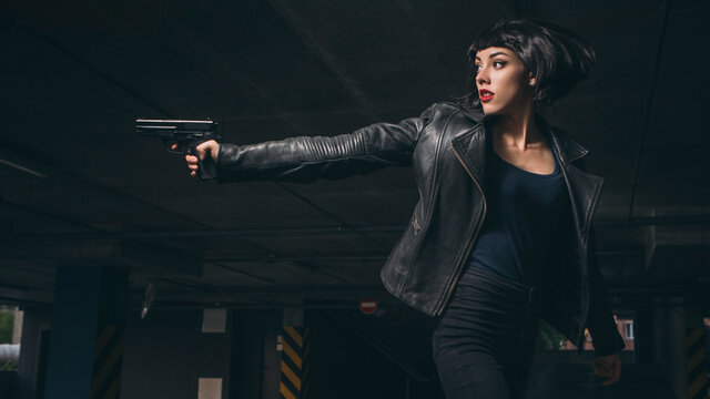Beautiful brunette sexy spy agent (killer or police) woman in leather jacket and jeans with a gun in her hand running after someone, to catch him on parking