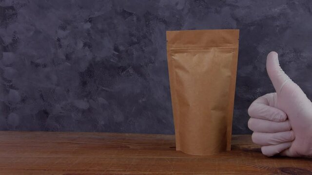 Hand in rubber gloves holds brown paper pouch bags mockup with coffee beans shows thumb up like wooden background Packaging food delivery goods template.Kraft pack with clasp tea leaves weight product