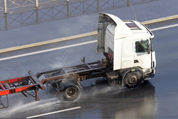 Truck with trailer and an empty orange long platform driving on wet asphalt after rain.