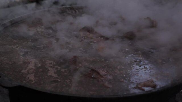 The meat is cooked in a cauldron. Night time. A slow fire burns and steam comes out of the cauldron.