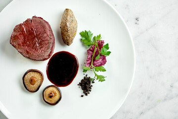 Appetizing medium rare beef steak with mushrooms and berry sauce, served in a white plate on a marble background. Restaurant food