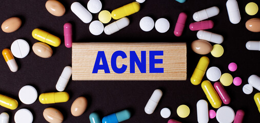 On a dark background, multi-colored pills and the word ACNE on a wooden block. Medical concept