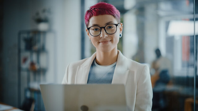 Modern Office: Portrait of Beautiful Authentic Specialist with Short Pink Hair Standing, Holding Laptop Computer, Looking at Camera, Smiling Charmingly. Working on Design, Data Analysis, Plan Strategy