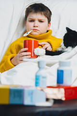 Sick child boy with digital thermometer in mouth lying in bed with fever illness headache stays home. Cute Kid with black cat in cozy sweater drinking herbal tea on couch taking temperature. Health.