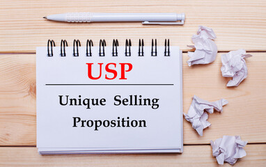 On a wooden background, a white notebook with the inscription USP Unique Selling Proposition, a white pen and crumpled white pieces of paper