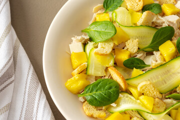 Naklejka na ściany i meble Plate with salad close-up on a brown background, salad of cucumber and chicken breast, mozzarella and croutons with sweet mango pieces and spinach