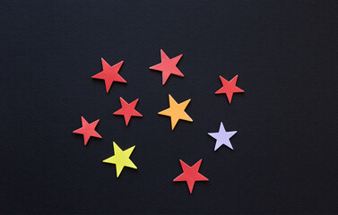Many colorful paper stars of various sizes randomly scattered on dark background; star color design; top view, flat lay