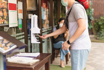 Man in medical mask using automatic alcohol gel dispenser spraying on his hand.