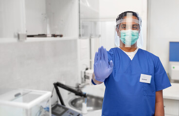 Fototapeta na wymiar medicine, healthcare and protection concept - indian doctor or male nurse in blue uniform, protective medical mask and face shield showing stop gesture over laboratory or hospital background