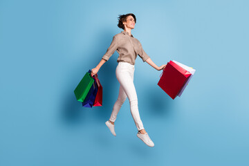 Full size profile portrait of cheerful pretty lady jumping hands hold bags isolated on blue color background