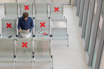 Obraz premium Male tourist sitting on row seats in airport lobby chair with social distancing sign and using laptop notebook computer. New normal journey concept