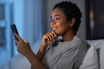 technology, internet, communication and people concept - happy smiling young african american woman in glasses with smartphone lying in bed at home at night