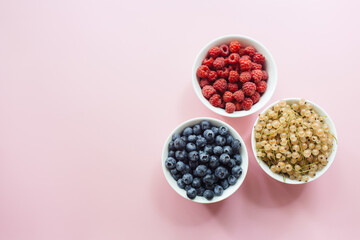 Variety of fresh currant berries white, blueberries, raspberries in white bowls on pastel pink background. Flat lay, top view, copy space