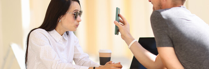 Naklejka premium Woman in sunglasses sit at round table and look in surprise at phone screen. Man show woman picture on phone.