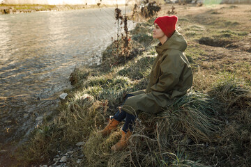 A young tourist girl in a raincoat, hat and tourist boots sits on the grass by the river Sunny weather in a journey Contemplation of the surrounding nature