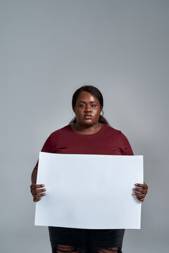 Serious plus size young african american woman in casual clothes looking at camera, holding blank white banner in front of her, posing isolated over gray background
