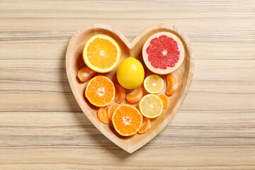 Many fresh citrus fruits in heart shaped box on wooden background, top view