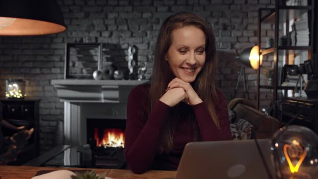 Home office - woman working from home with laptop computer. Talking on video chat, sitting at desk in dark living room in front of fireplace in Winter.