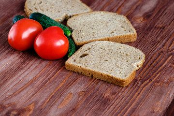 Two heart-shaped tomatoes lie on a wooden table along with bread and gherkins. Copy space