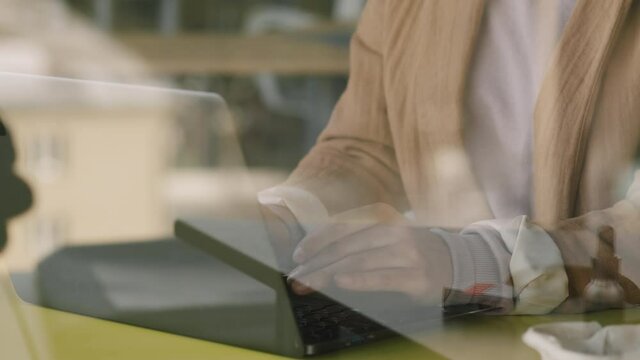 Close up mid-section with PAN of hands of unrecognizable businesswoman sitting behind glass wall or clear protective screen and typing on laptop