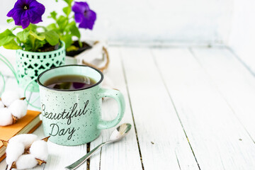 Close-up of a cup of tea on a wooden white table with blurred background, front blur. Still life with flower, book, teaspoon, cotton box. Spring breakfast. Copy space.