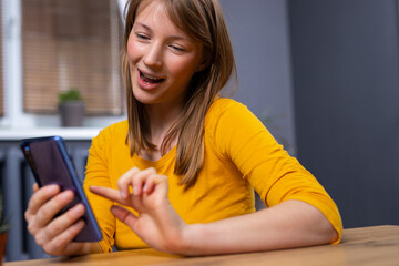 Image of happy nice, blonde, young woman in pajamas, smiling and using cellphone while sitting on the floor in living room