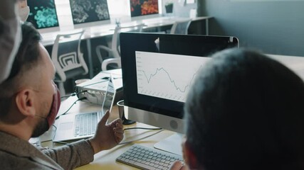 Handheld slowmo over the shoulder shot of businesspeople in face masks looking at line chart on computer screen and discussing work in office amidst pandemic - Powered by Adobe