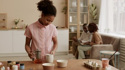 Locked-down shot of happy African elementary-aged girl pouring tea from teapot into mug, bringing it to mother, sitting with father on couch at home, people smiling