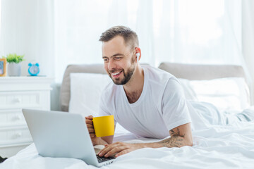 Portrait of a smiling young bearded man lying on bed in the morning and working distantly on his...