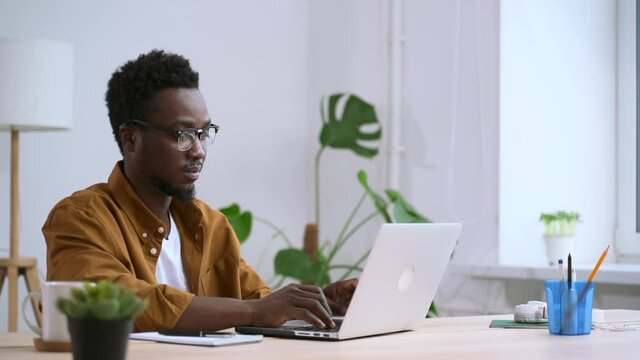 African man in glasses on laptop works in white home interior background of window Spbas. Male model with beard works on computer in comfortable environment. Room has green plant and lamp for lighting