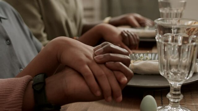 Side view of three cropped unrecognizable people holding hands on top of table, praying before dinner