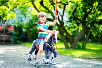 Little kid boy playing with his toy rocking horse in a summer park. Happy smiling child having fun outdoors. Blond boy of 5 years