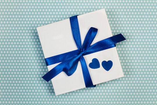 Caja de regalo blanca con un lazo azul junto a corazones azules sobre un fondo celeste con motivos. Vista superior. Copy space