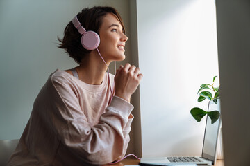 Happy beautiful girl in headphones working with laptop at window
