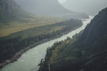 Misty mountain landscape with wide mountain river. Dark green gloomy scenery with confluence of two...