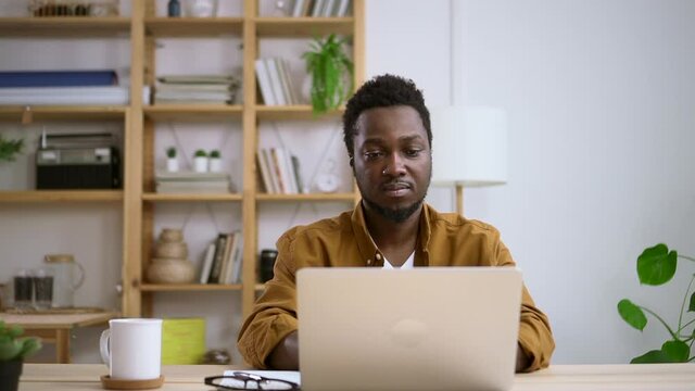 African man closes laptop lid after finishing work on home interior background Spbas. Black leather male model types on computer keyboard and then exits and logs out. Person smile at table behind