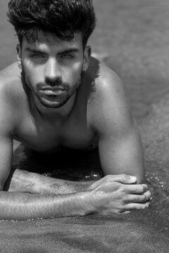 Retrato de chico hispano joven y guapo tirado en la arena. Fotograf&iacute;a en blanco y negro, primer plano. Hombre con barba y mirada seductora