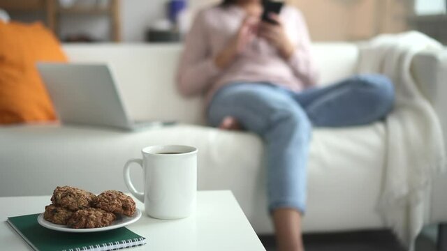 Closeup of coffee cup and cookie, woman browse phone on defocused background Spbd. Sweet snack on table. Blurred female use mobile smartphone in cozy home on couch. Concept living room, bokeh