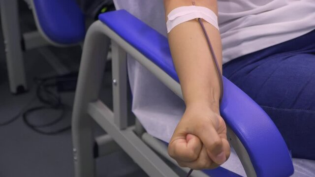 Front close-up, panning shot of a woman arm while opening and closing her hand while donating blood with a catheter attached with medical tape.