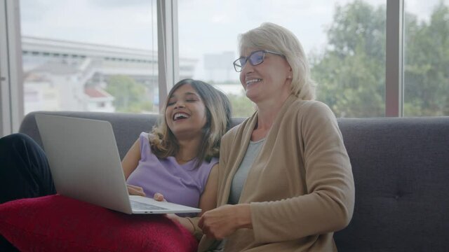 Medium shot - Adopted Asian daughter and her Caucasian stepmother enjoy watching movie on internet at home. Love and caring time of Multi-racial family concept.