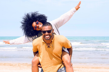 African american happy couple having fun piggyback riding and playing airplane on the beach - Black man and afro hair woman in playful moment on the shore at summer time - Concept of happiness image - Powered by Adobe