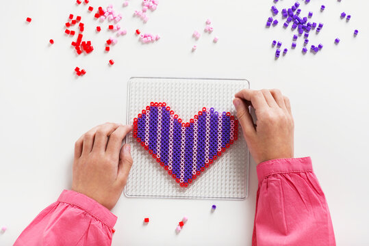 Little child hands make beads art in the shape of heart. Children education concept. Kids crafts. Top view.