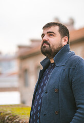 Bearded guy in dark blue sweater leaning on a stone wall in the street, looking at the sky seriously.