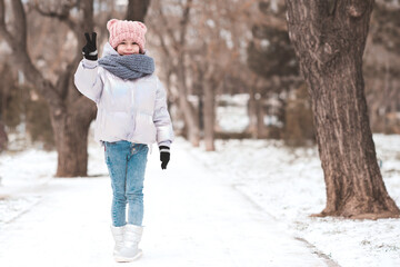 Happy kid girl 5-6 year old having fun with snow in park. Wearing stylish winter clothes: knitted hat, gloves and scarf, boots and jaket. Happipness.