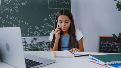 schoolgirl talking on smartphone and looking at notebook near laptop on desk