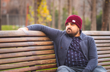 Boarded guy in a woolen hat and sweater sitting on park bench worried.