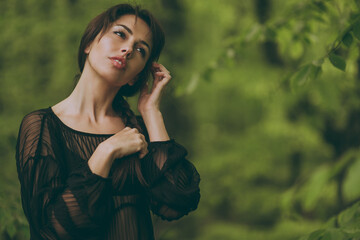 Beautiful young woman in a black transparent chiffon dress among the green foliage of forest trees