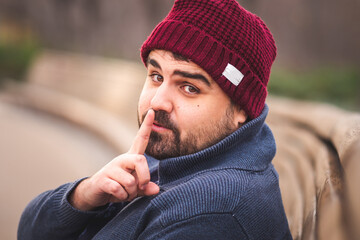 Boarded guy in a woolen hat and sweater sitting on park bench. His finger on his mouth (asking for silent) and looking at the camera. 