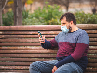 Guy with protective mask using his smartphone on a park bench at sunset. 